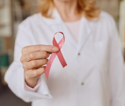 Warm-toned close up of unrecognizable female doctor holding pink ribbon as symbol of breast cancer awareness, copy space